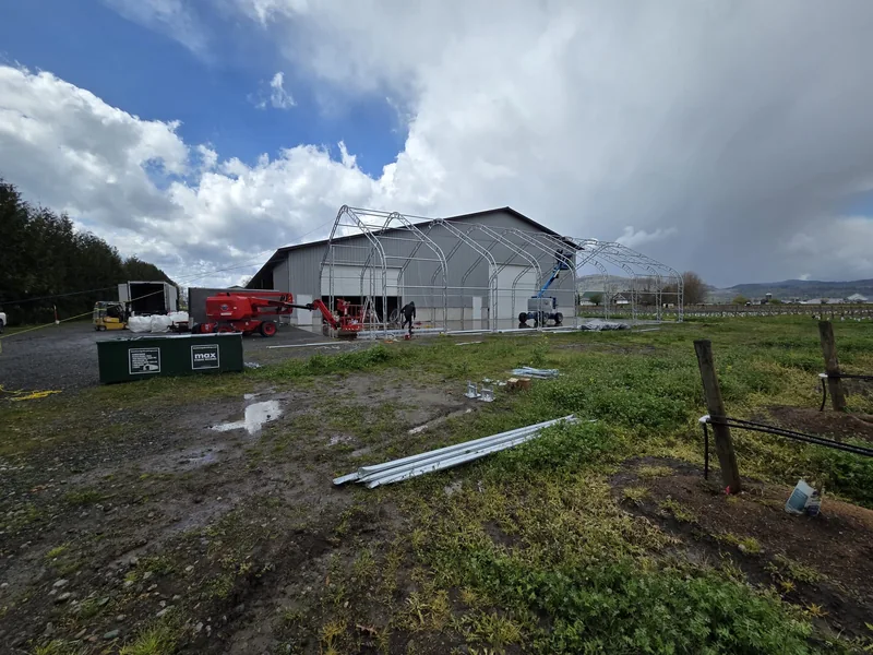 Steel arches under assembly beside an existing building and rows of crops